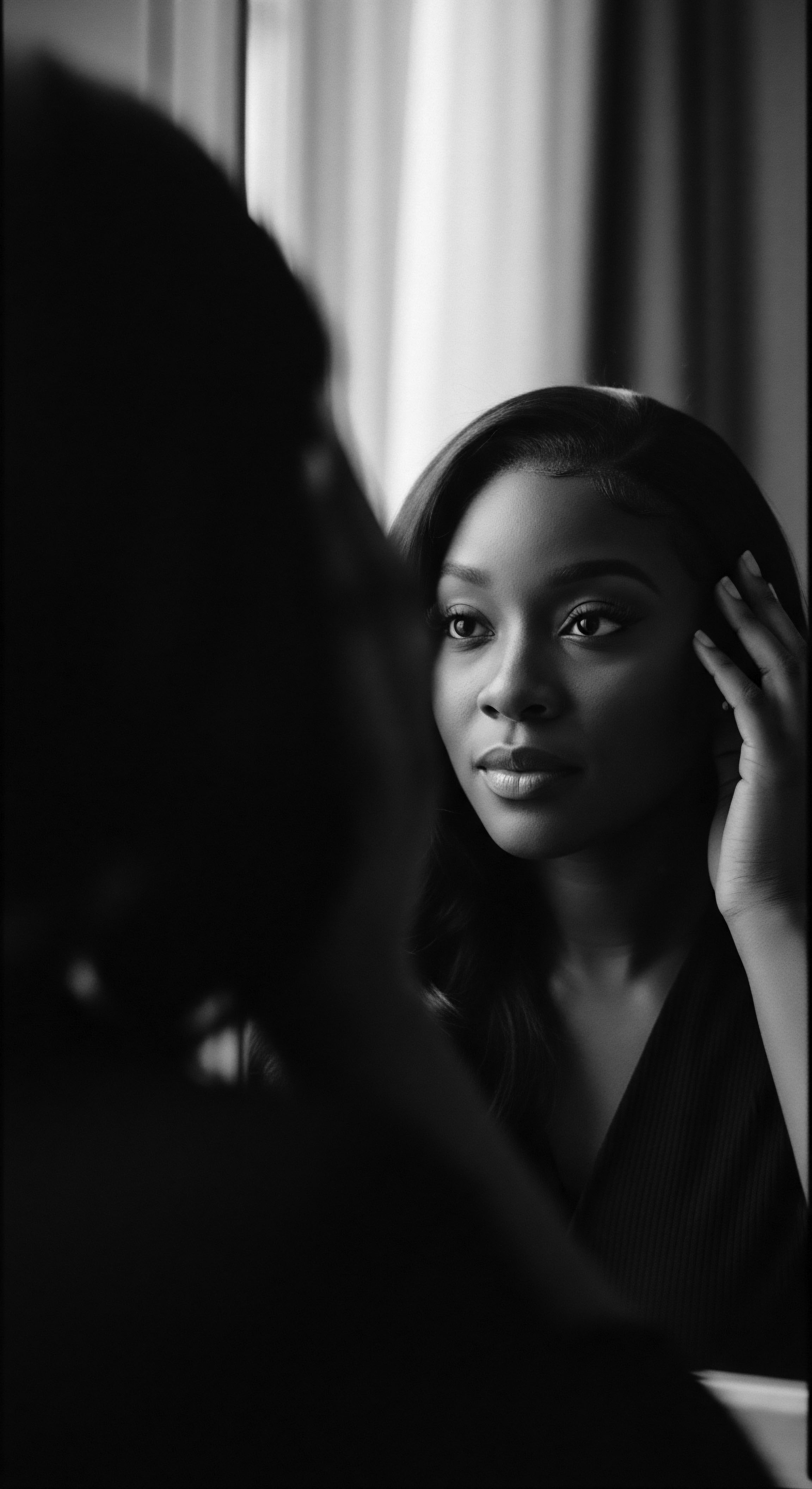 A poised woman is seen in monochrome, her coiled hair cascading softly around her face, framed by her delicate hand. The reflection introduces depth, highlighting her contemplative expression. Textured hair care is intrinsically linked with heritage and cultural expression. The holistic approach to hair underscores the celebration of its unique beauty.