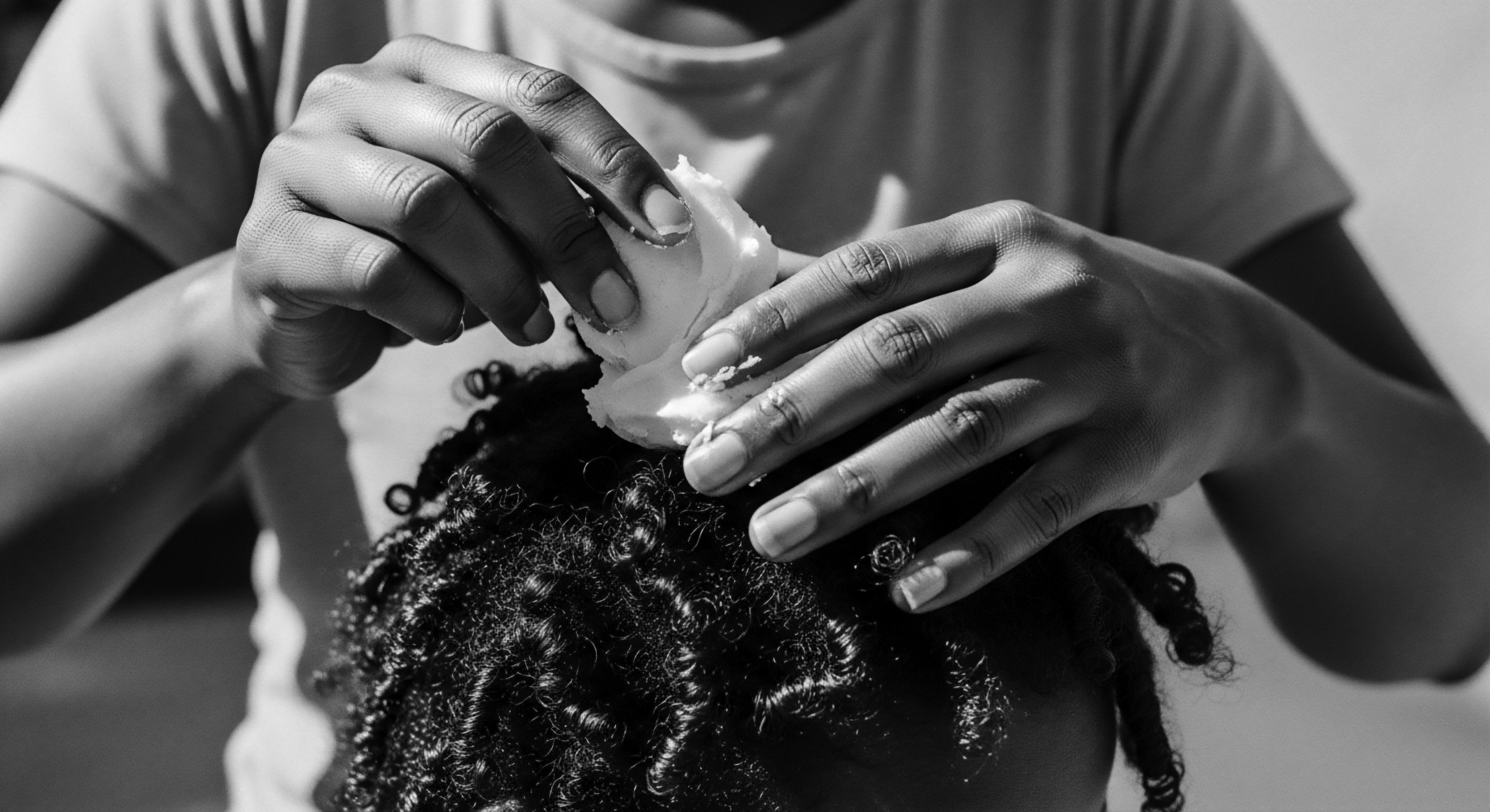 A close-up in monochrome captures hands gently applying a creamy emollient to densely coiled hair. The fingers work methodically, ensuring even distribution of the hydrating treatment designed to enhance the natural texture and resilience rooted in ancestral heritage with wellness and identity. Light and shadow emphasize the hair's intricate formations.